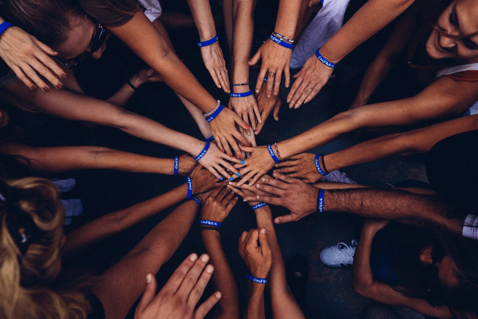 A bird’s eye view of a team standing in a huddle, with their arms reached out and their hands meeting in the center, as teams often do before a game. Each team member is wearing a blue bracelet. 